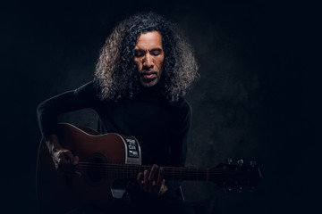 Fototapeta premium Handsome man with long hair is playing acoustic guitar at dark studio.