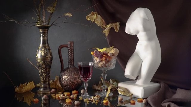 Still life with a plaster bust of Venus on a background of drapery, branches with autumn leaves in a vase, dried fruits and red wine