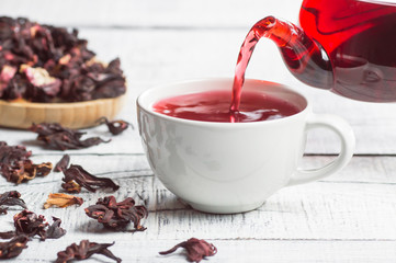 White cup of healthy hibiscus tea pouring from the teapot with dried hibiscus flowers on white wooden background, winter hot drink concept for cold and flu 