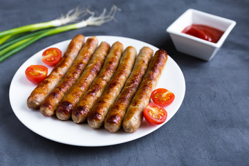 Grilled, delicious sausages lie on a white plate on a gray background. Next to the plate are chopped cherry tomatoes, behind the plate is an onion and is the sauce in a gravy boat.