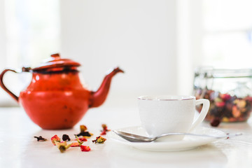 View of cup of tea, red teapot, herbal and infusion elements in a white room. Breakfast, food, and wellness concept. 