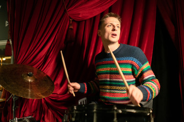 handsome young guy playing drums on stage. against the background of the red curtain