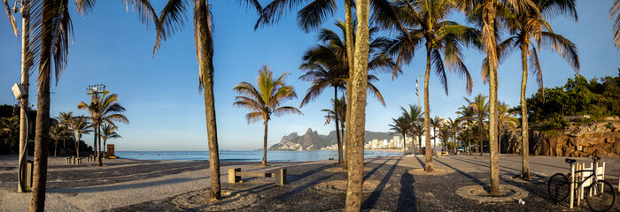 Wide panorama from the Arpoador boulevard showing the sunrise over Ipanema beach in Rio de Janeiro with  several palm trees in the foreground and the Two Brothers mountain in the background
