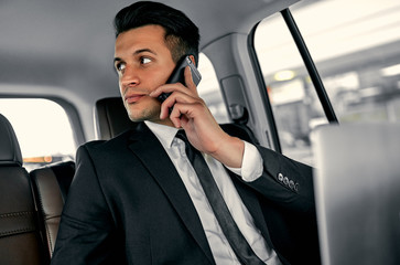 Young handsome businessman is sitting in luxury car. Serious handsome man in suit is working with laptop and talking on smart phone while being in trip.