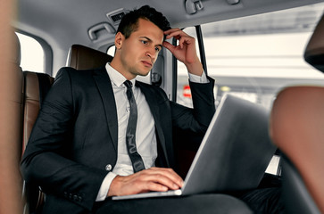 Young handsome businessman is sitting in luxury car. Serious man in suit is working with laptop while being in trip.