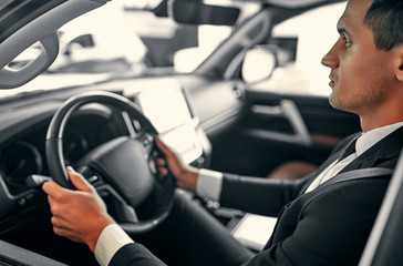 Young handsome businessman is sitting in luxury car. Serious man in suit is driving.