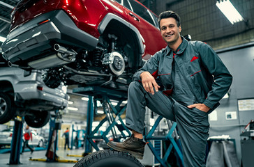 Handsome mechanic in uniform is working in auto service. Car repair and maintenance. Holding car wheel. - obrazy, fototapety, plakaty