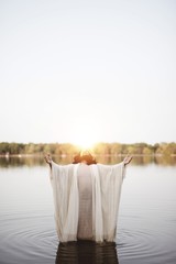 Vertical shot of the Jesus Christ standing in the water with his hands and head towards the sky