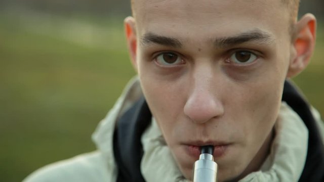 Pensive man smoking electronic cigarette and exhale clounds of vapor in the park at chilly late autumn evening, close up