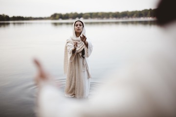 Female wearing a biblical gown while standing in the water and reaching towards Jesus Christ