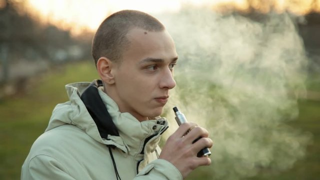 Pensive man smoking electronic cigarette and exhale clounds of vapor in the park at chilly late autumn evening, close up