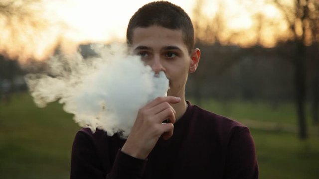 Relaxed caucasian young man with short hair vaping e-cigarette in the park in the evening, Close up