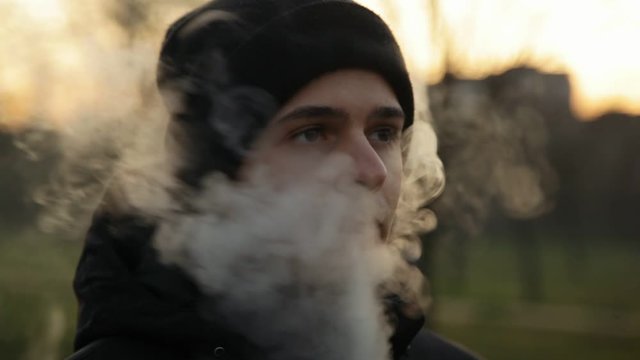 Pensive man smoking electronic cigarette and exhale clounds of vapor in the park at chilly late autumn evening, close up