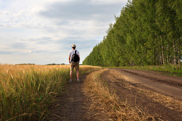 young man traveler with backpack in hat stands in field at sunset and looks away, concept of freedom of choice, future, concept of travel and adventure