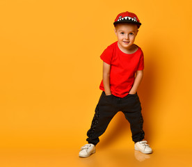 Young guy boy in a red T-shirt and dark pants, white sneakers and a funny cap posing on a free copy space on a yellow background