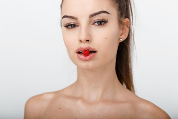 Portrait of a young girl with ponytail, with naked shoulders, holds in the mouth a raspberry, isolated on a white background. Concept about youth, young skin, eyebrows, eyelashes, beauty