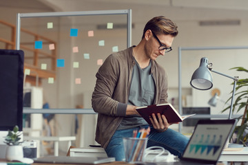 Side view portrait of handsome adult businessman sitting on desk in office and reading note book pensively, copy space