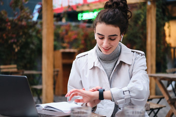 Pretty smiling brunette girl in trench coat happily using smartwatch studying in cafe on city street