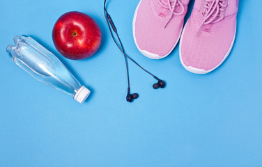 Pink sport shoes, earphones, red apple and bottle of water on a blue background. Concept healthy lifestyle, sport and diet. 