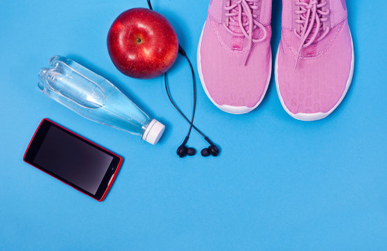 Pink sport shoes, earphones, red apple, smartphone and bottle of water on a blie background. Concept healthy lifestyle, sport and diet. © Snowbelle