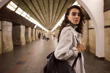 Beautiful girl in trench coat with backpack confidently looking in camera at subway station
