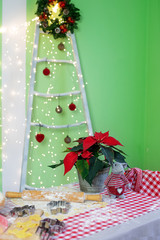 A table on which lies dough, flour and cookie cutters for Christmas cookies, in a room decorated for Christmas or New Year. On the table sits a gray mouse and there is a poinsettia