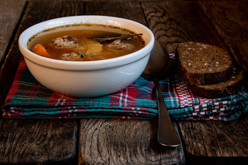 Homemade soup with meatballs in a white toe, on a kitchen checkered napkin with bread and grains and a teaspoon.