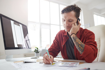 Portrait of contemporary tattooed businessman speaking by phone and taking notes while working at...