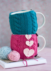 Two cups in pink and blue sweater stand one on one. One of the cups has hearts of felt. Nearby are two tangles of knitting threads and an old notebook.