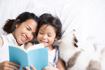 Mother and daughter read a book in her bed. And there is a dog lying down.