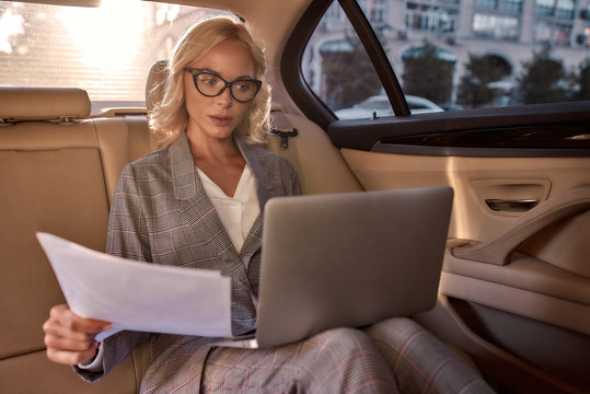 Preparing for a business meeting. Beautiful and successful woman in full suit analyzing documents and working on laptop while sitting in the ca