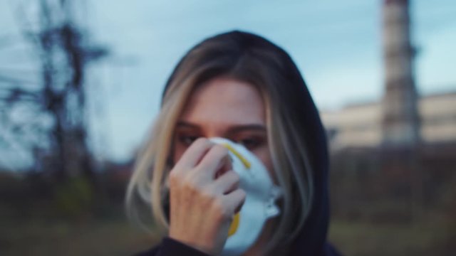 Young female person wearing breahte protection face mask holding poster slogan Climate Crisis Ahead. Private factory with emissions tower on background. Day light. Global politics. Ecology involution.