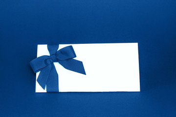 Isolated white gift card with trendy blue colored ribbon bow on blue background. Color of the year 2020 concept. Holiday greeting card concept. Copy space.