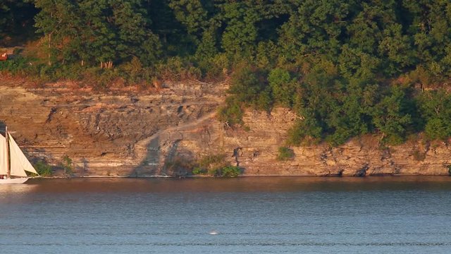 A two-masted schooner sailing ship passes a rocky cliff on a lake near sunset on Seneca lake, one of the Finger Lakes of New York
