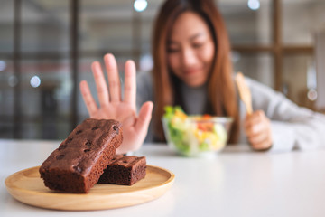 A woman choosing to eat vegetables salad and making hand sign to refuse a brownie cake on the table