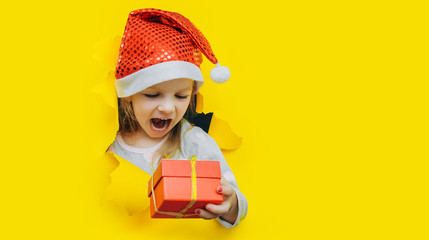 A cute little surprised child in a red Santa hat looks out of a hole in yellow paper and holds a box with a gift in her hand. Christmas and New Year concept. Bright background, copy space.