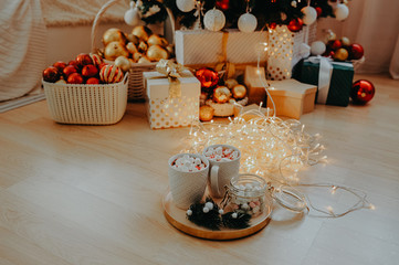 two mugs with a drink and marshmallows, on a gold tray with a can of marshmallows and a sprig of a Christmas tree, against the background of a Christmas tree in the lights and gifts under it