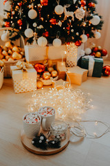 two mugs with a drink and marshmallows, on a gold tray with a can of marshmallows and a sprig of a Christmas tree, against the background of a Christmas tree in the lights and gifts under it