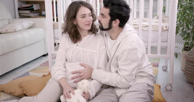 Man and Pregnant Woman Sitting on Floor, Embracing and Laughing. Young Family Expecting a Baby. White Interior with Baby Cot on the background. Concept of Parenthood, Happiness and Tenderness