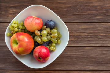 Fruits apples, grapes, plums in white plate on wooden background. Cooking, recipe, diet, healthy food concept. Top view, copy space