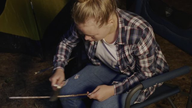 Tourist girl scraping wooden stick with knife in campsite for evening picnic. Girl using knife for scrabing wooden stick for barbeque in forest hike at dark evening