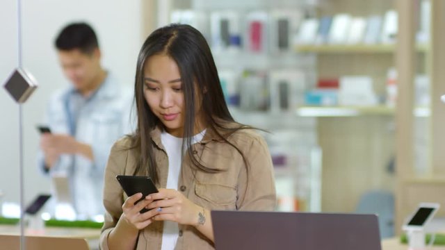Tracking shot medium shot of young Asian woman standing at gadget store and using smartphone