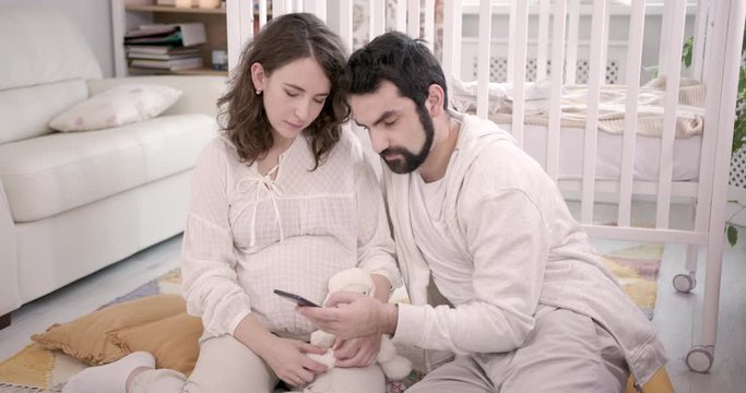 Man and Pregnant Woman Sitting on Floor, Use Mobile Phone, Embracing and Laughing. Young Family Expecting a Baby. Baby Cot on the Background. Concept of Parenthood, Happiness and Tenderness. 