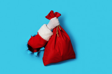 Santa Claus hand holding red sack full of presents over light blue