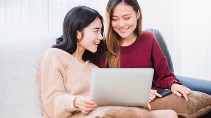 Fototapeta premium Young beautiful Asian women lesbian couple lover using smartphone and laptop video call online in living room at home with smiling face.Concept of LGBT sexuality with happy lifestyle together.