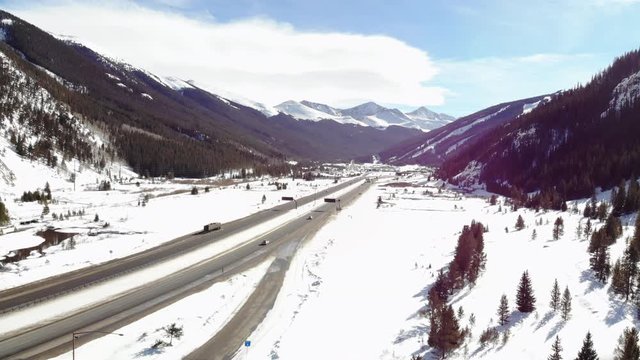 Drone shot of a slow pan from left to right over a snow covered Colorado highway with snow top covered mountains in the distance and pine tree covered mountains to the left and right of highway