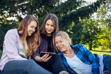 Three young pretty women on the lawn in park look at smartphone and laugh. Female students relax in the park sitting on green grass. Blonde, brunette and brown-haired outdoor, close-up.