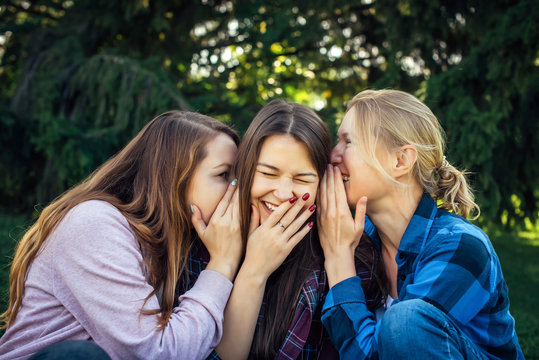 Three young attractive woman sharing secrets sitting on green grass in the park. Cheerful girlfriends gossip and whisper outdoor.