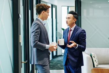 Serious Vietnamese entrepreneur with cup of coffee discussing gossips with colleague during break in office corridor