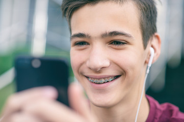 Close-up portrait of a smiling young man with a smartphone, in the street.  Happy teenage boy is using mobile phone, outdoors. Cheerful teenager spends time in social networks using cell phone.